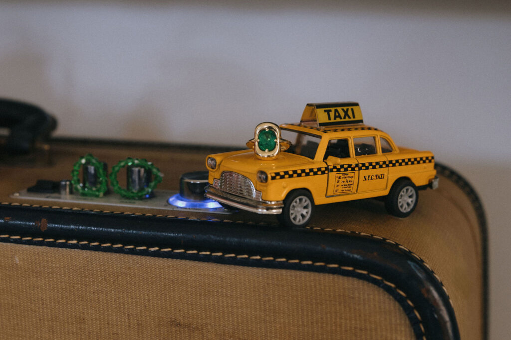 Close-up of a miniature yellow NYC taxi adorned with a ring featuring a green gemstone, set atop a vintage amplifier with jade bracelets in the background