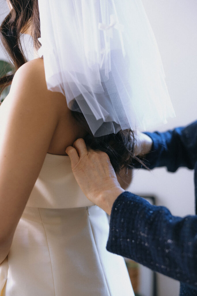 Close-up of a mother adjusting the back of her daughter's strapless wedding gown, with a delicate veil adorned with bows resting gently over her hair.