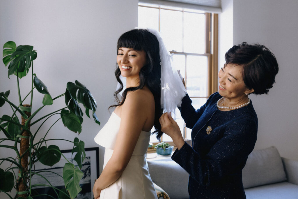 A radiant bride smiles as her mother adjusts her delicate veil adorned with tiny bows, with soft natural light streaming through a nearby window.