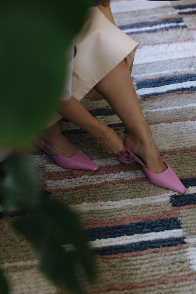 A bride slipping on vibrant pink pointed-toe slingback heels, captured on a textured striped rug with a hint of greenery in the foreground.