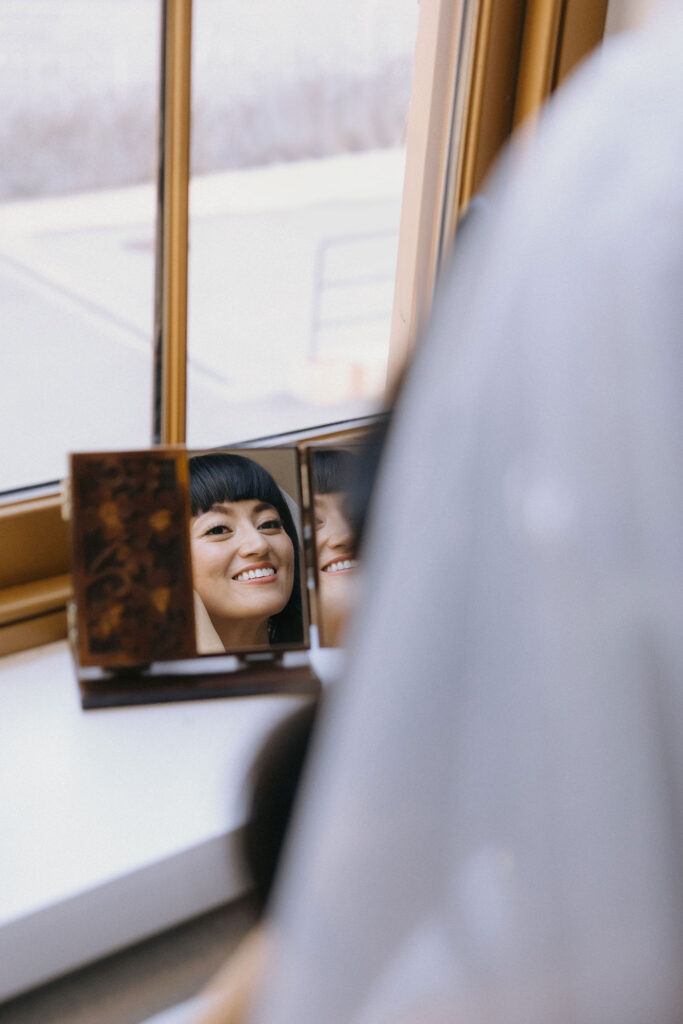 A bride’s radiant smile reflects in a vintage hand mirror placed by a sunlit window, capturing her excitement and beauty on her special day