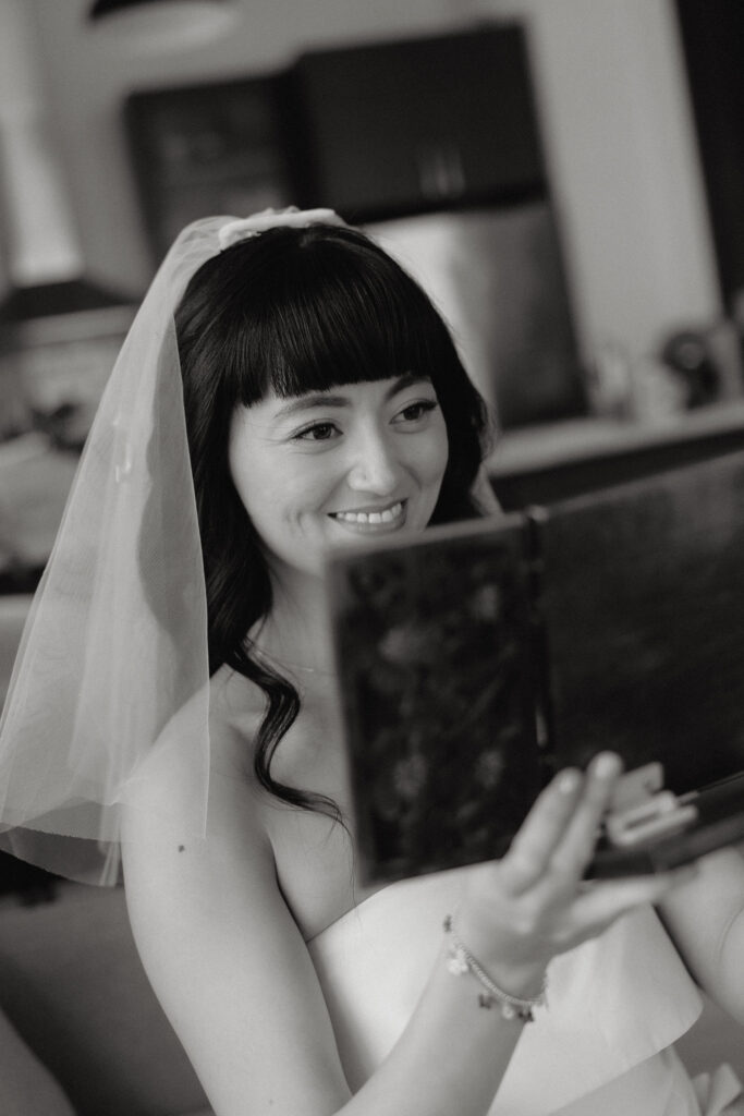 A bride with soft curls and a simple veil smiles warmly while holding a mirror, her joy reflecting in her eyes as she admires herself. The intimate black-and-white portrait captures her natural beauty and serene excitement.