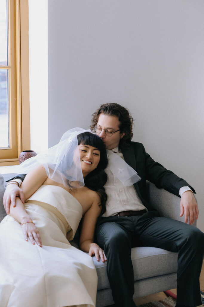 A bride reclines on a cozy couch, leaning into her groom who tenderly kisses her head. The bride wears a minimalist strapless gown and a delicate veil with bow details, while the groom dons a tailored dark suit with glasses, exuding quiet intimacy.