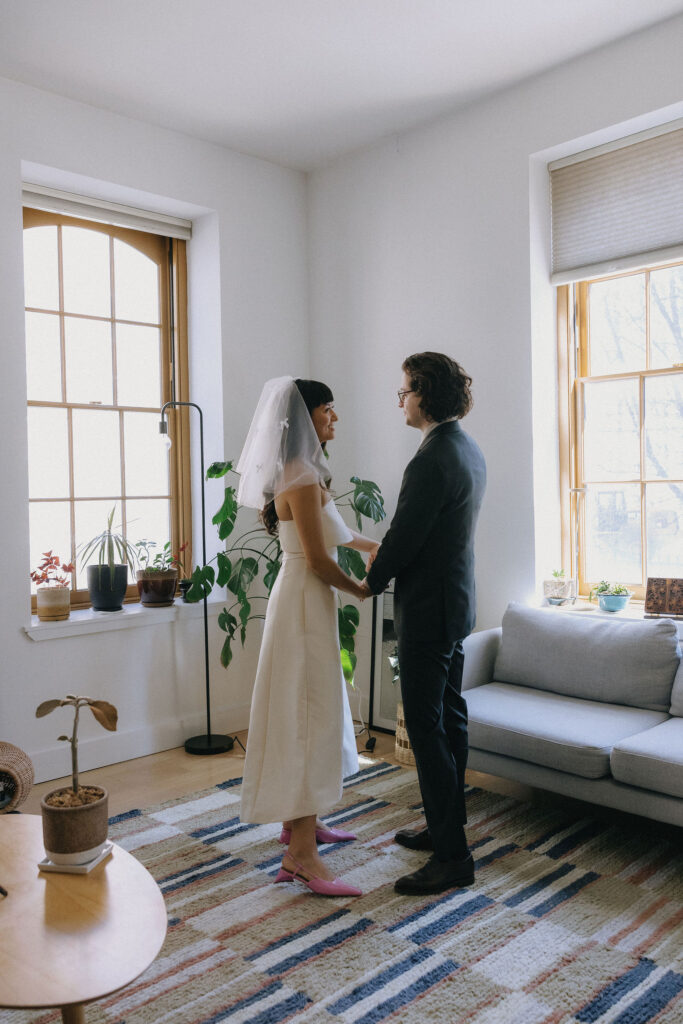 A bride and groom stand together in a serene and sunlit living room, holding hands and sharing a loving gaze. The bride wears a cream dress, veil with bows, and pink heels, while the groom looks dapper in a dark suit. Houseplants and soft furnishings add a cozy charm to the setting.