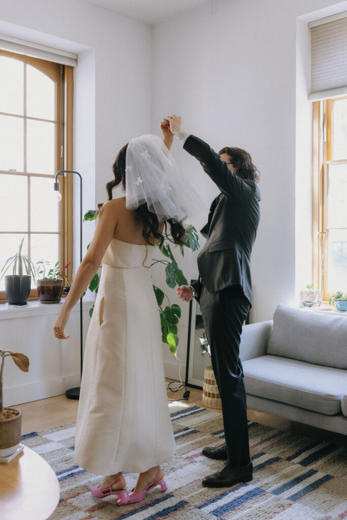 A romantic moment as the groom spins his bride in a sunlit room, showcasing her elegant cream dress and veil with bow details. The bride wears pink heels, adding a playful touch to the intimate scene surrounded by cozy home decor.