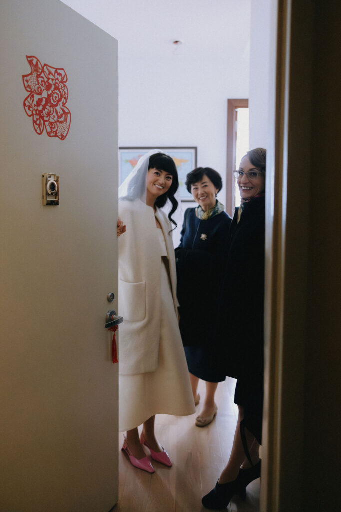 A candid photo of a smiling bride in a white coat and veil standing by an open door, accompanied by two women in elegant attire. A red decorative symbol adorns the door, adding a cultural touch to the scene