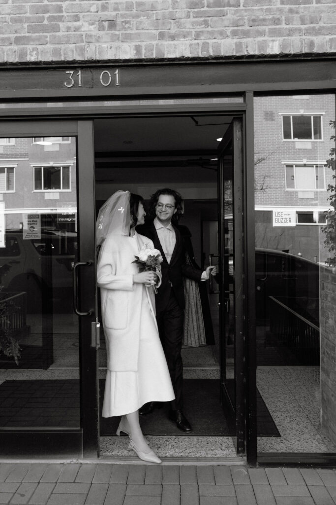 A black-and-white photo of a bride and groom stepping out of a building, smiling at each other. The bride is in a simple, elegant white coat and veil, holding a bouquet, while the groom wears a dark suit and glasses, holding the door open