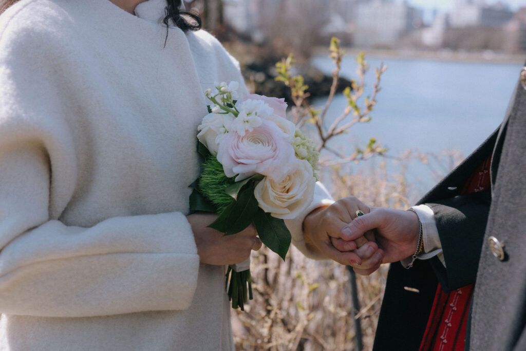 A bride in a soft white coat holding a bouquet of pale pink and white flowers with lush greenery, gently clasping her partner's hand adorned with a green-stone ring against a serene riverside background.