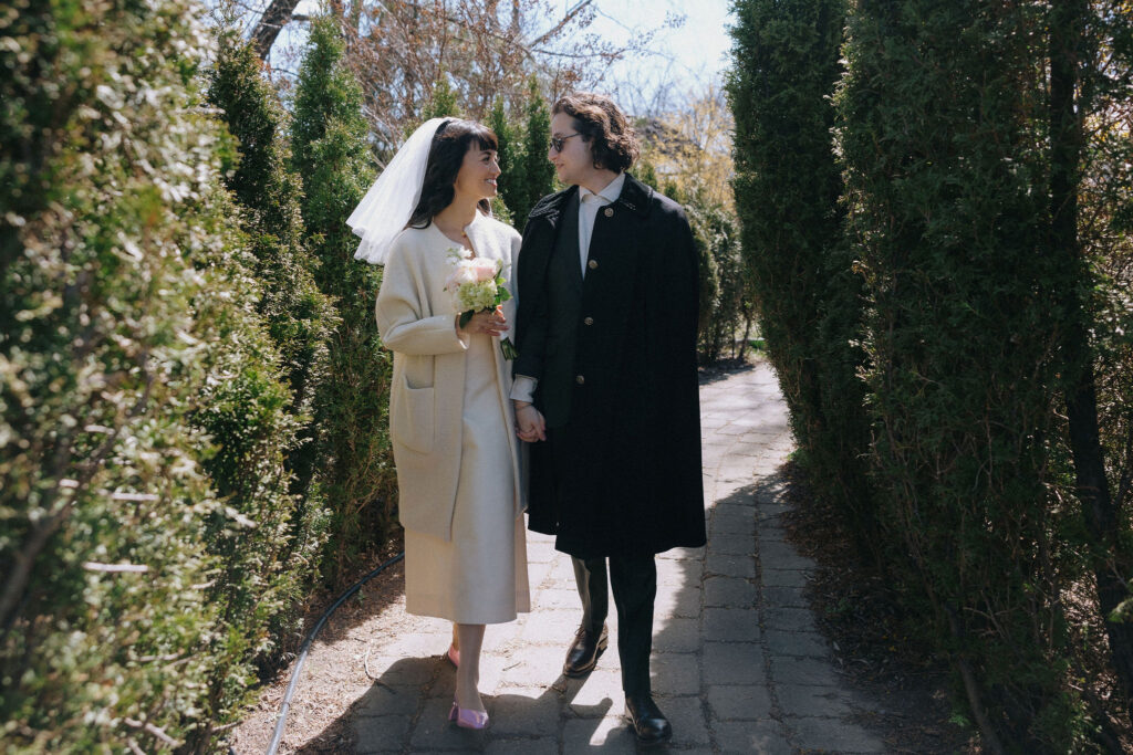 An elated couple walking hand-in-hand through a sunlit garden path, surrounded by lush greenery. The bride, wearing a veil and holding a bouquet, gazes lovingly at her groom, who is dressed in a formal black coat.