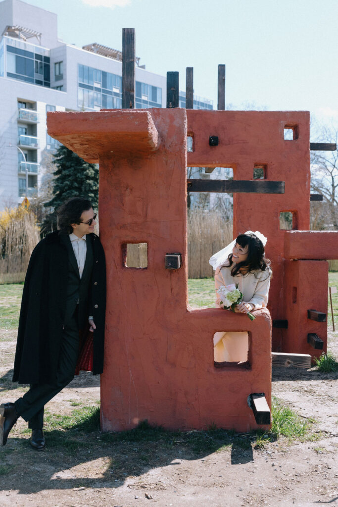 A playful couple poses around a rustic red structure in an outdoor urban park, with the bride holding a bouquet and the groom leaning on the wall, both smiling.