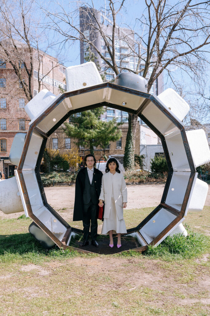 An elegant couple stands within a geometric outdoor art installation in NYC, framed by urban architecture and a clear blue sky