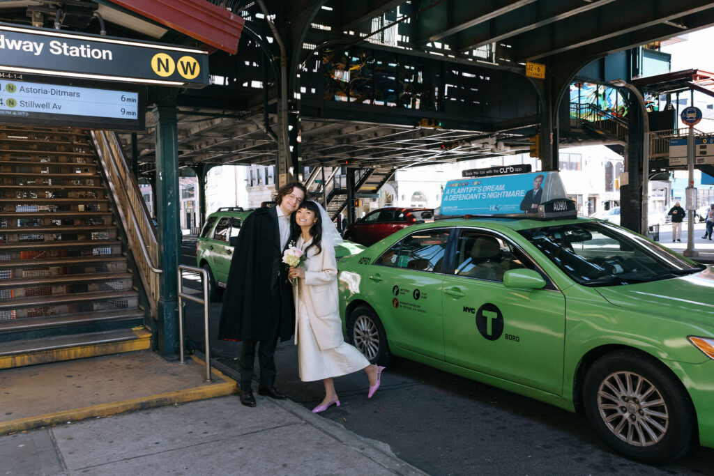 A newlywed couple poses at a subway station entrance under the elevated tracks in NYC, with a green taxi nearby, capturing the essence of a modern city elopement