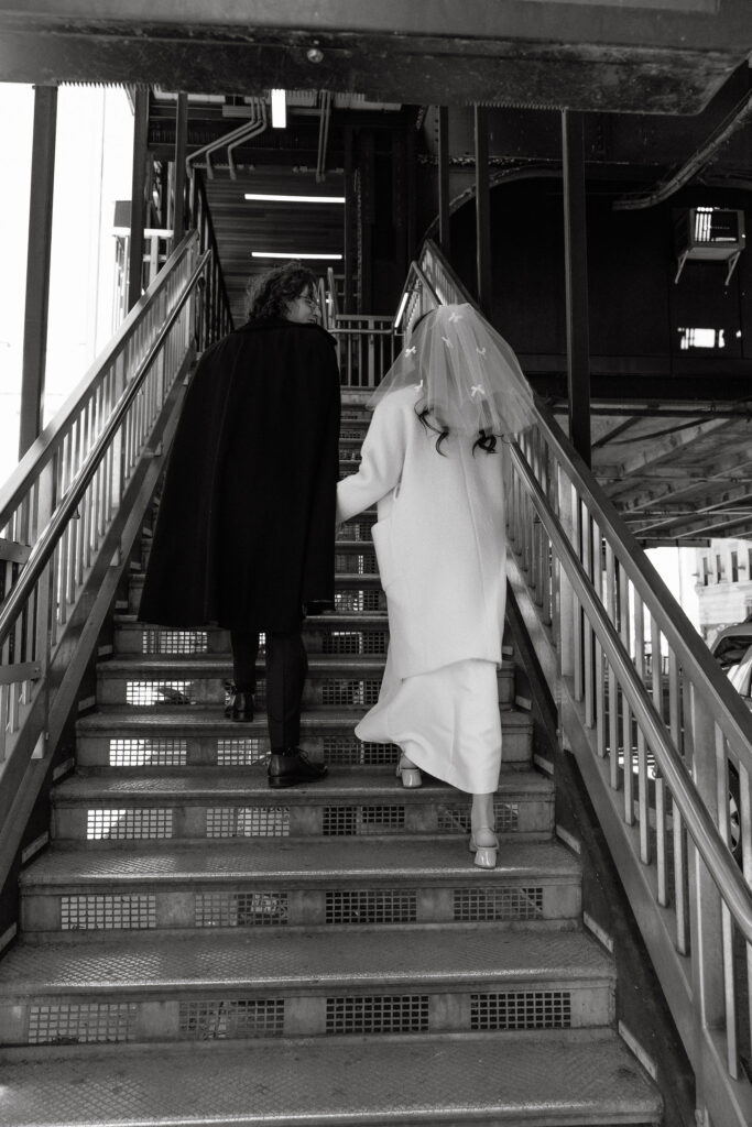 Black-and-white photo of a bride in a veil with bows walking up a staircase with her partner, surrounded by urban architecture