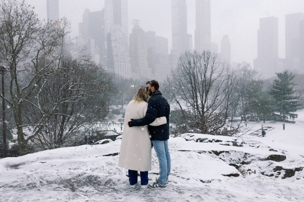 Couple enjoying the winter NYC skyline view, with him kissing her hair in the snow before proposing