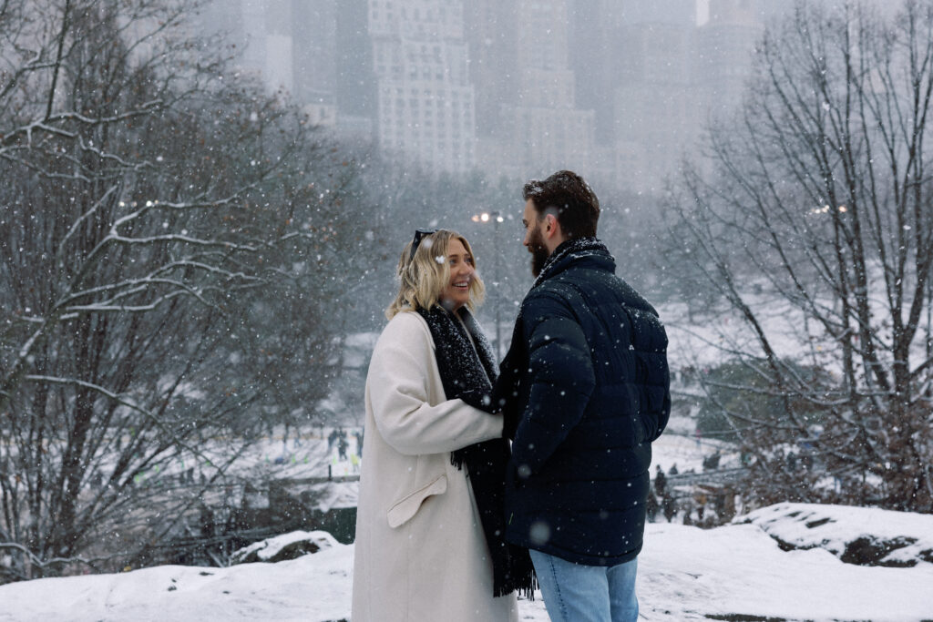 Moments before a proposal in snow-covered Central Park, a smiling woman radiates happiness and anticipation in a romantic setting