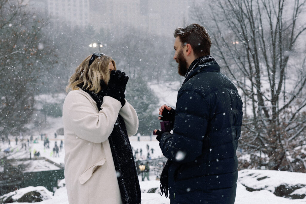 Surprised woman in the snow as she realizes her partner is proposing with a ring, hands raised to her face in shock and joy.