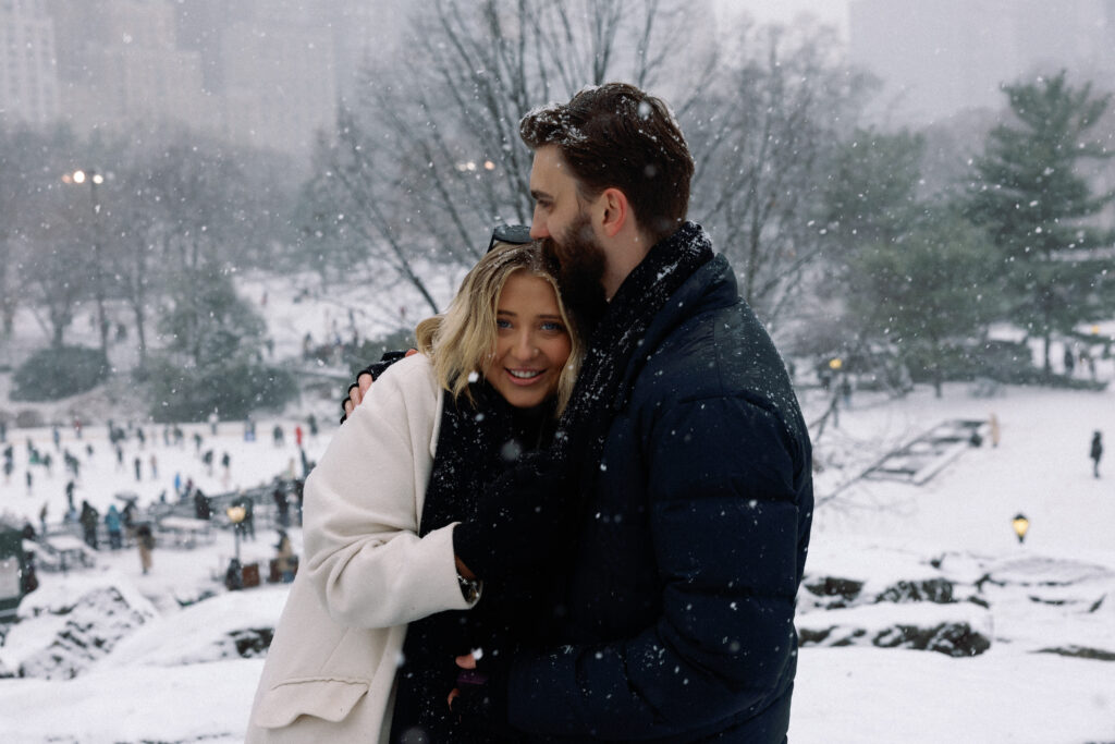 Couple right after their proposal, with her resting on his chest as he kisses her hair in a snowy setting