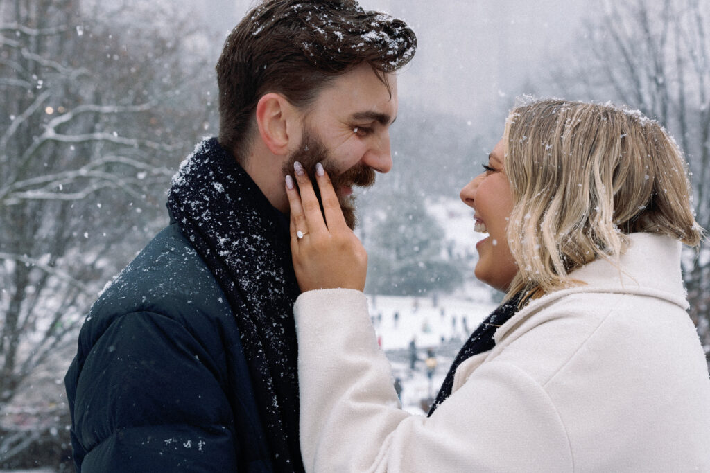 Couple laughing and looking at each other as she gently touches his face, showcasing her engagement ring in a romantic and joyful moment