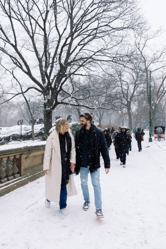 Newly engaged couple laughing and walking through the snow in Central Park, radiating joy and love.