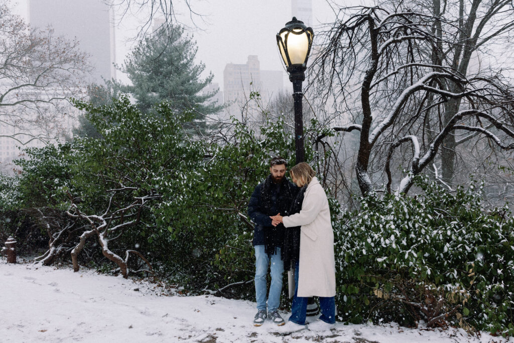 Couple admiring an engagement ring by a lightstand in Central Park, with snow-covered greenery in the background