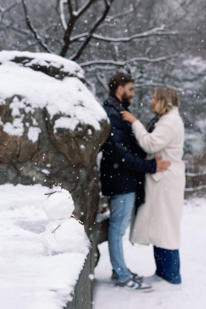 A snowman in focus with a couple out of focus in the snowy background of Central Park