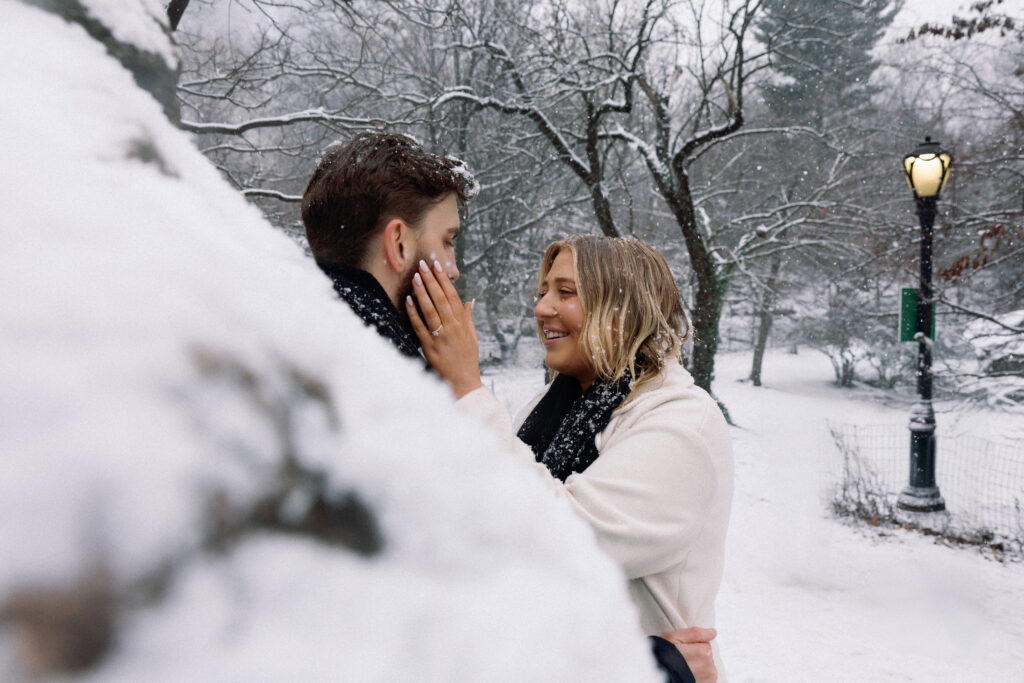 Fiancée gently places her hand with an engagement ring on her partner's cheek during a snowy moment in Central Park