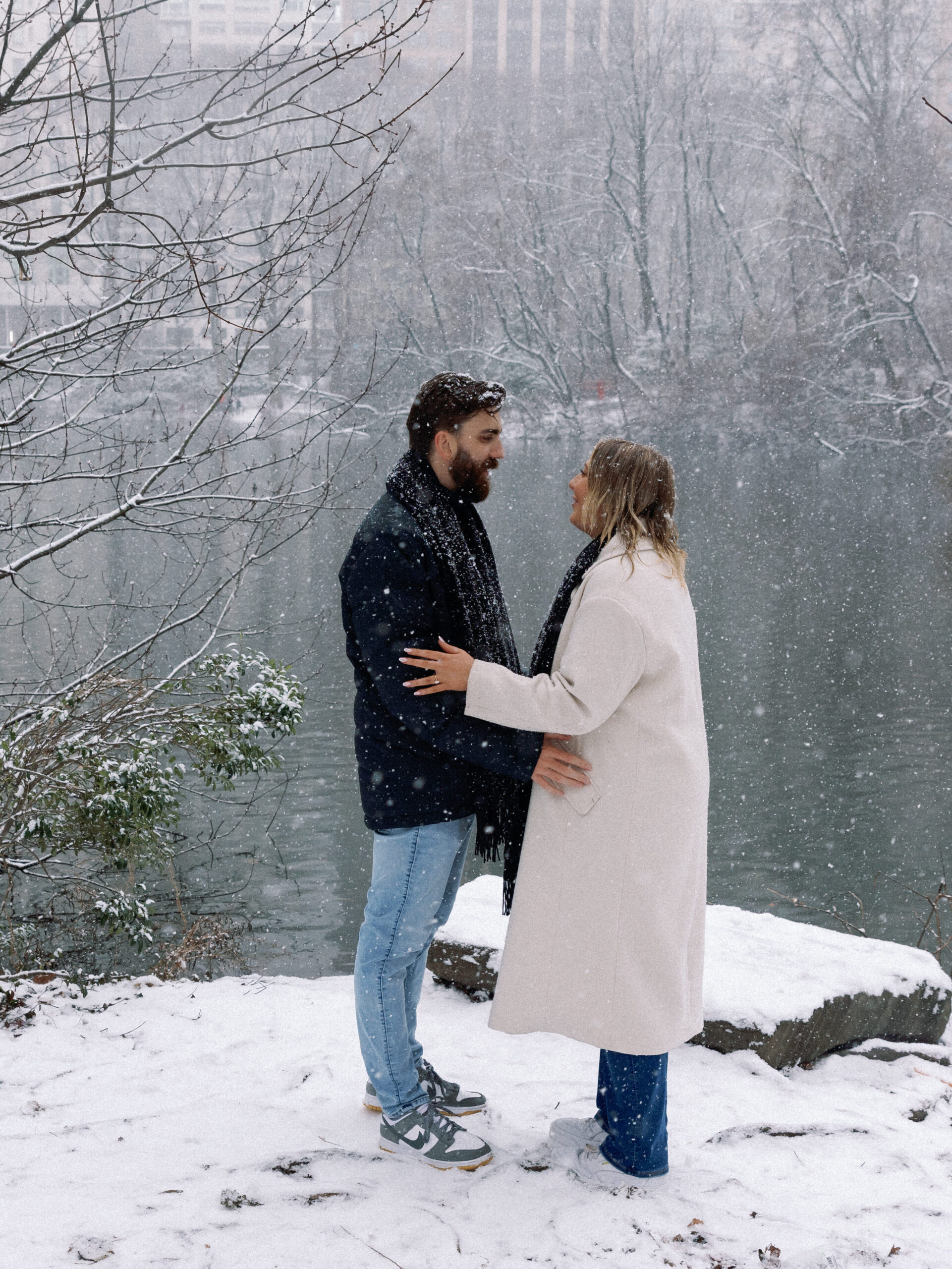 Couple standing by the pond under Gapstow Bridge in Central Park, surrounded by snow in a romantic winter setting.