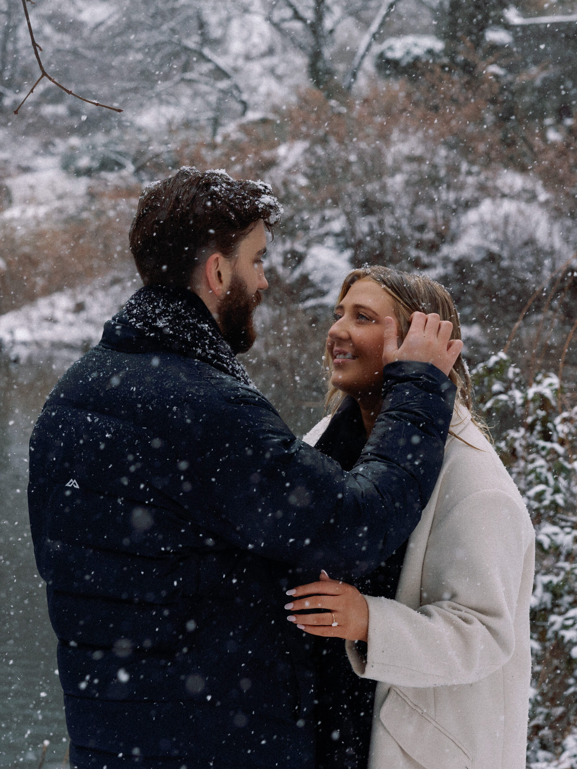 After a snowy proposal in Central Park, a fiancé gently brushes snow from his partner’s hair in a tender moment.