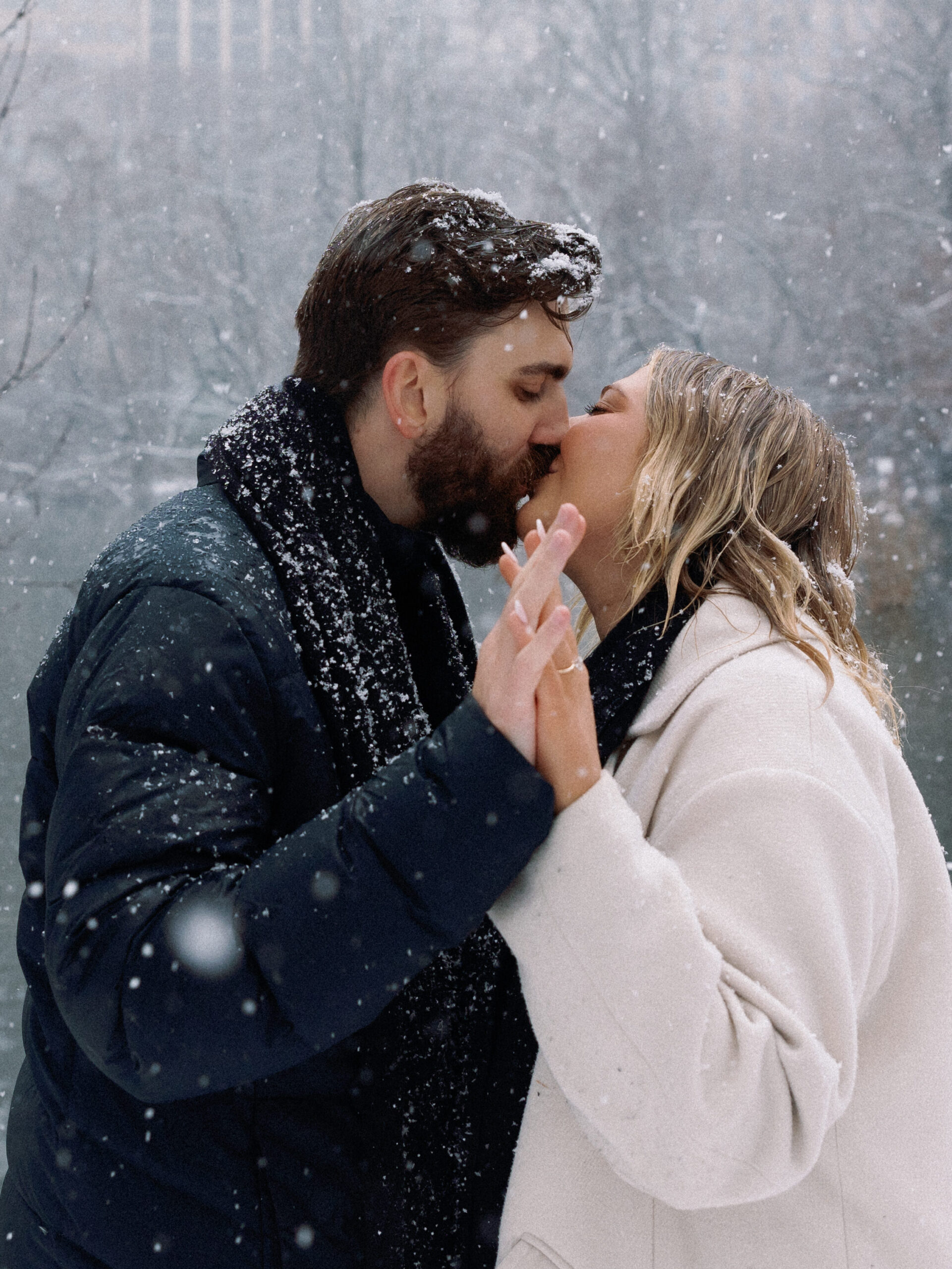 Romantic kiss in snowy Central Park with intertwined hands and an engagement ring visible.