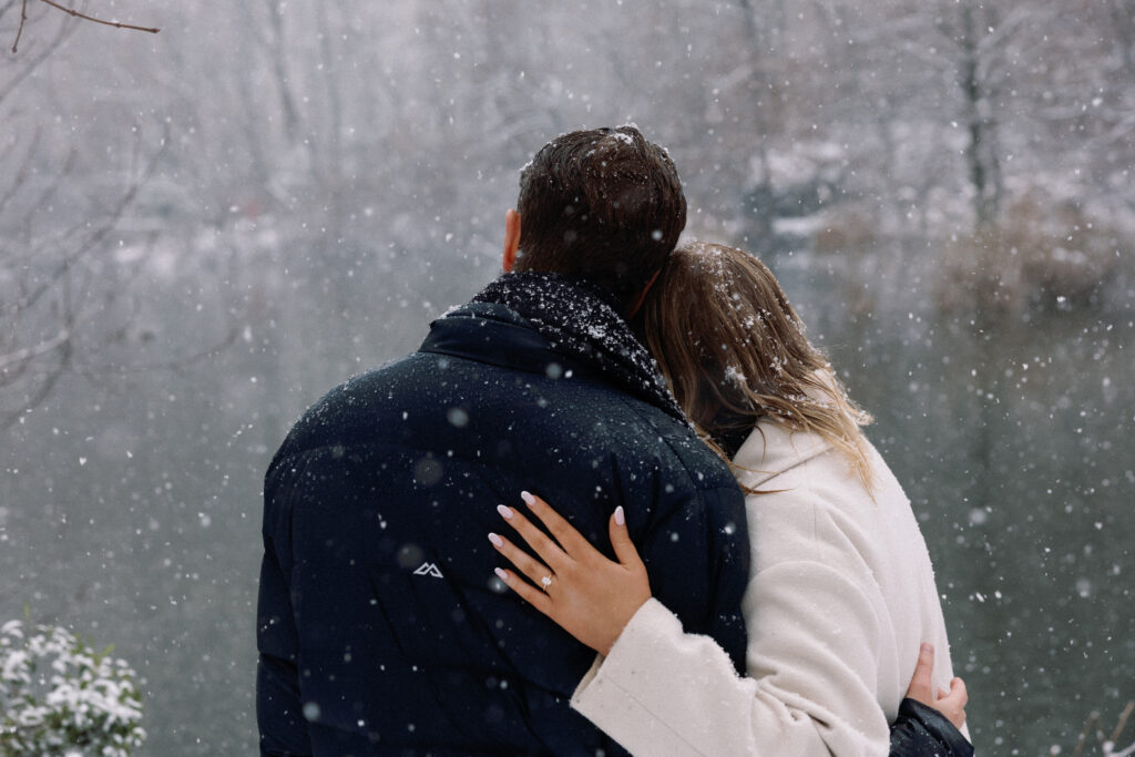 Back view of a couple in Central Park, heads tilted toward each other, with a snow-covered scene, water, and engagement ring visible