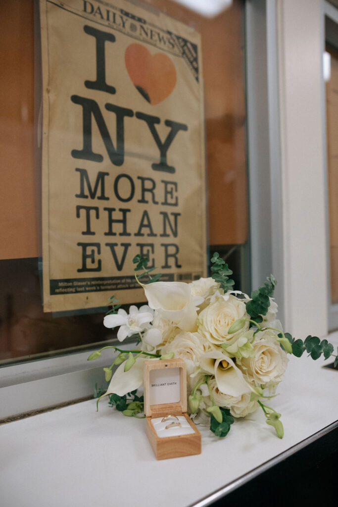 White rose bouquet and wedding rings displayed in a wooden box with an 'I ❤️ NY More Than Ever' sign in the background at Brooklyn City Hall