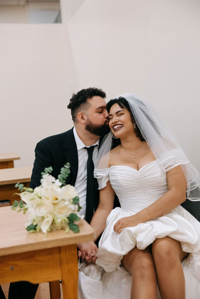 Groom kisses the bride on the cheek as they sit together holding hands with a white rose bouquet on the table at their Brooklyn City Hall elopement