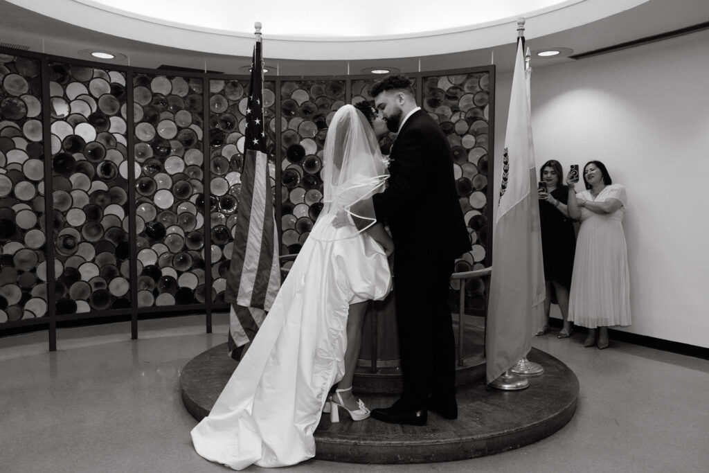 The bride and groom share their first kiss during their Brooklyn City Hall elopement ceremony, with loved ones cheering in the background