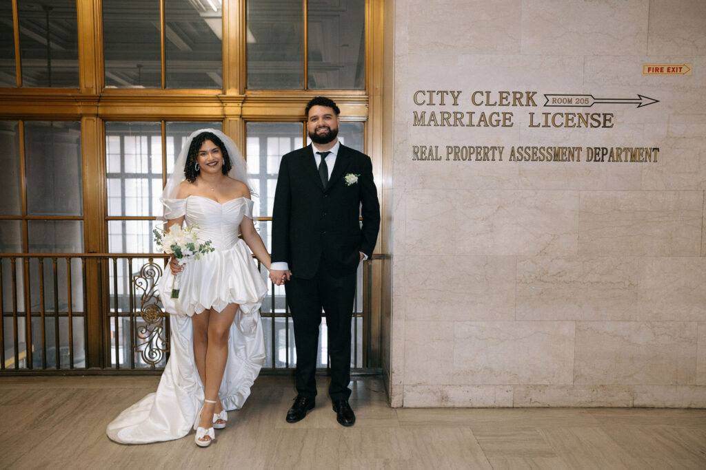 The newlywed couple stands smiling at Brooklyn City Hall, holding hands near the City Clerk Marriage License sign, with the bride in a modern wedding dress and bouquet.