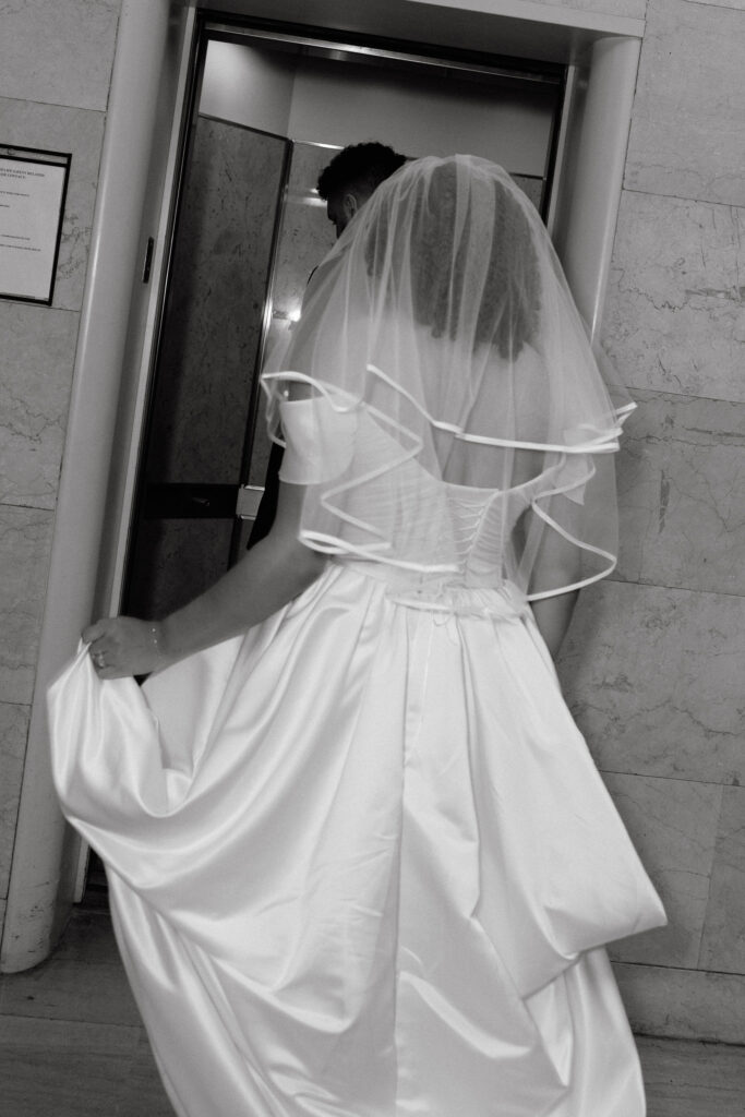 Bride in a flowing satin wedding gown with a veil walking into an elevator at Brooklyn City Hall, capturing a candid moment during her elopement