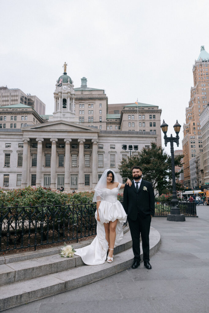 Bride and groom pose together on the steps in front of Brooklyn Borough Hall during their NYC elopement, with a bouquet resting nearby