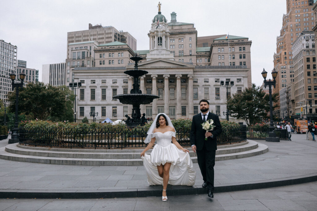 Bride and groom pose in front of the fountain at Brooklyn Borough Hall after their NYC elopement, with the historic building in the background