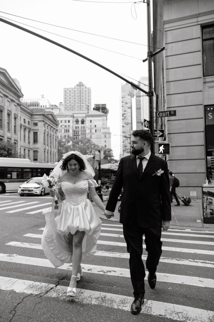 Bride and groom hold hands while crossing the street near Brooklyn Borough Hall during their NYC elopement, with the bride smiling joyfully