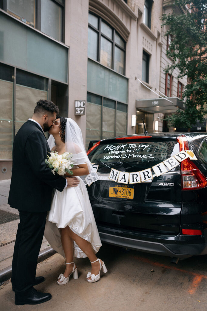 Bride and groom share a kiss next to their decorated car with a 'Just Married' sign after their Brooklyn City Hall elopement.