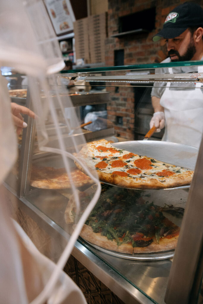 A close-up of fresh pizza slices on display at a Brooklyn pizza shop, with the bride’s veil visible in the foreground