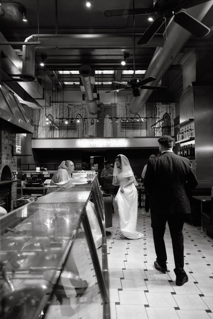 Bride and groom walk into a cozy Brooklyn pizza shop, enjoying a casual moment after their NYC elopement