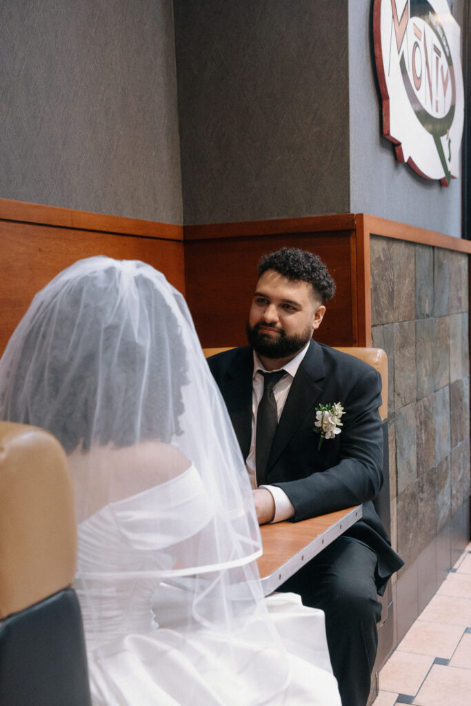 Groom gazes at his bride as they sit together in a booth at a Brooklyn pizza shop, celebrating their NYC elopement