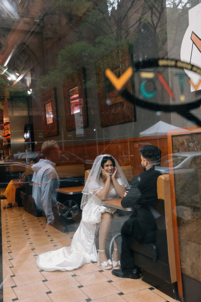 Bride and groom sharing a romantic moment at a Brooklyn pizza shop, captured through the shop window with reflections of the city