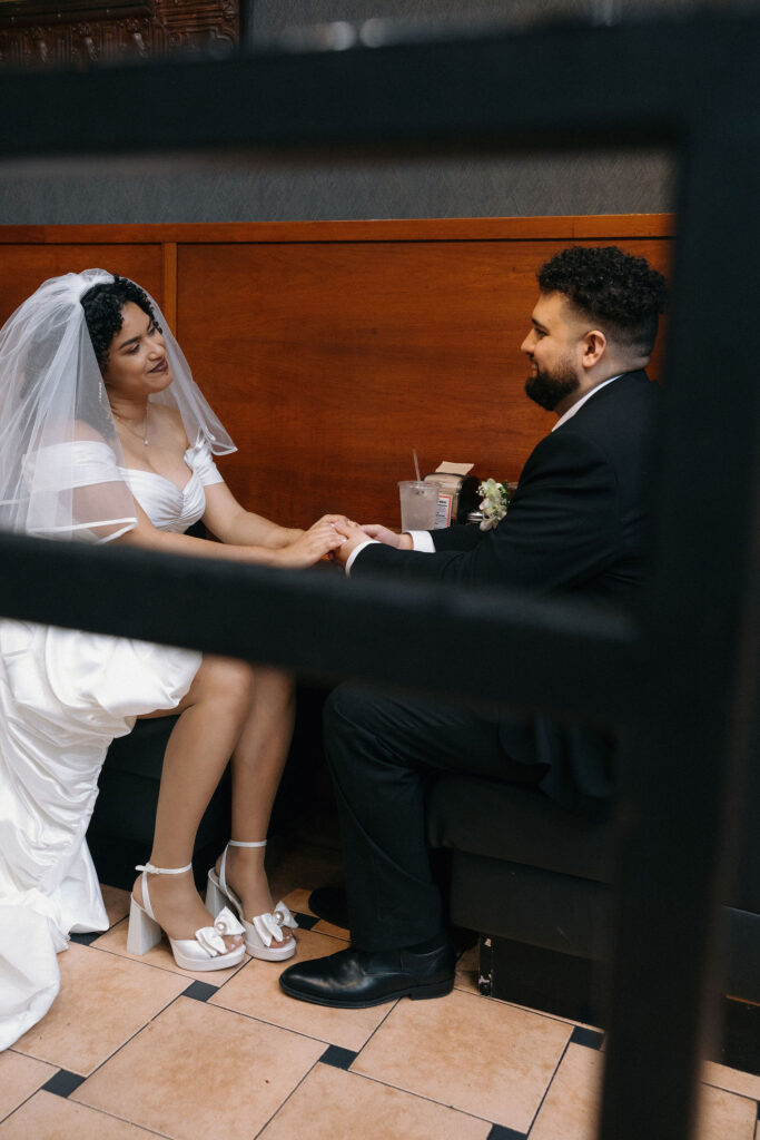 Bride and groom sitting at a booth in a Brooklyn pizza shop, holding hands and sharing a loving moment during their NYC elopement celebration