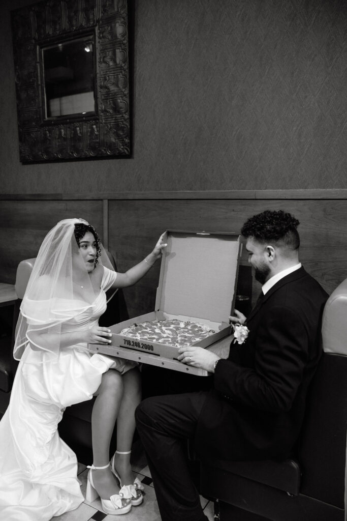 Bride and groom sharing a playful moment while opening a pizza box at a Brooklyn pizza shop after their NYC elopement.