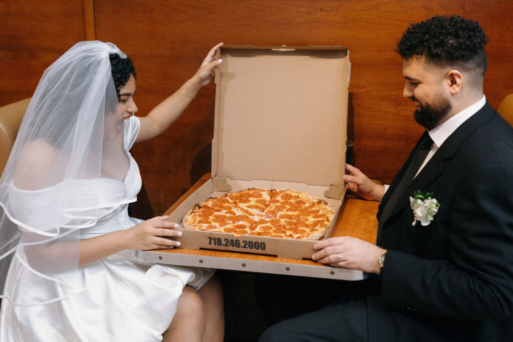 Bride and groom holding an open pizza box with a fresh pepperoni pizza, celebrating their NYC elopement in Brooklyn