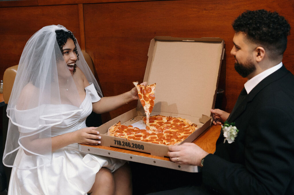 Bride happily holding a slice of pepperoni pizza from an open box while sharing a lighthearted moment with the groom at a Brooklyn pizza shop