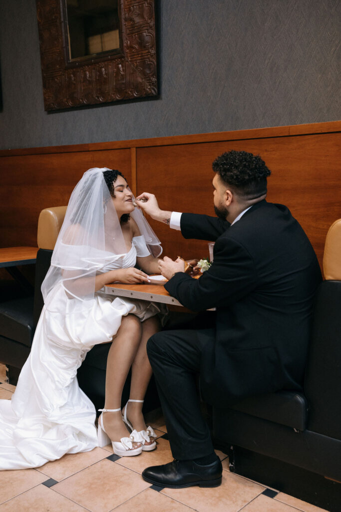 Groom feeding the bride a slice of pizza in a playful and romantic moment at a Brooklyn pizza shop during their NYC elopement.