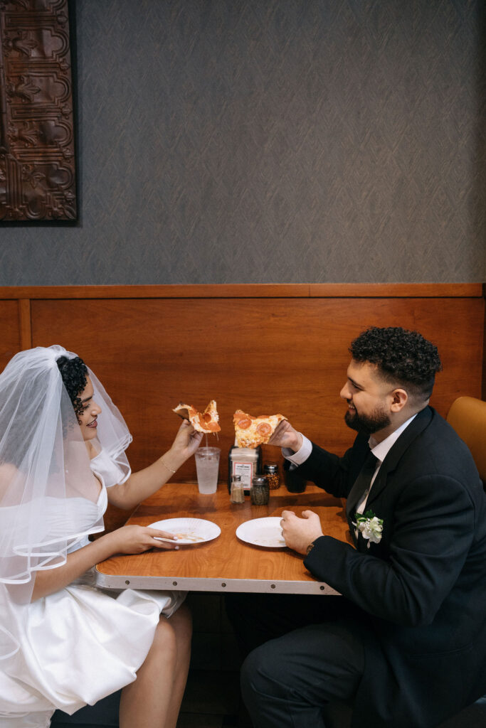 Bride and groom toasting with slices of pepperoni pizza at a Brooklyn pizza shop during their NYC elopement celebration