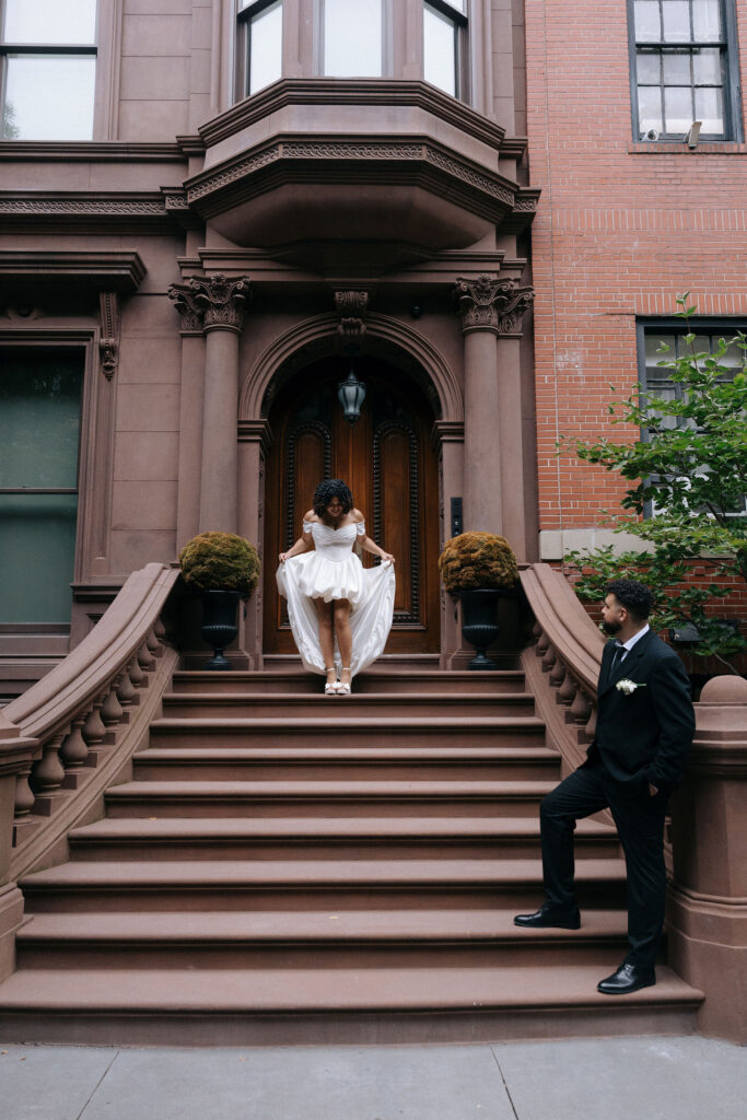 Bride standing on the steps of a classic Brooklyn brownstone while the groom admires her from below during their NYC elopement.